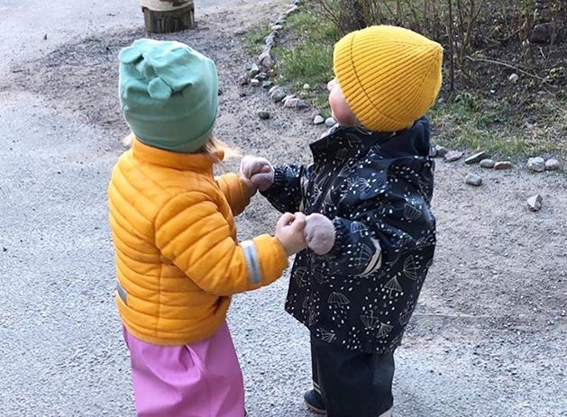 Två barn i regnkläder håller hand ute på gården.