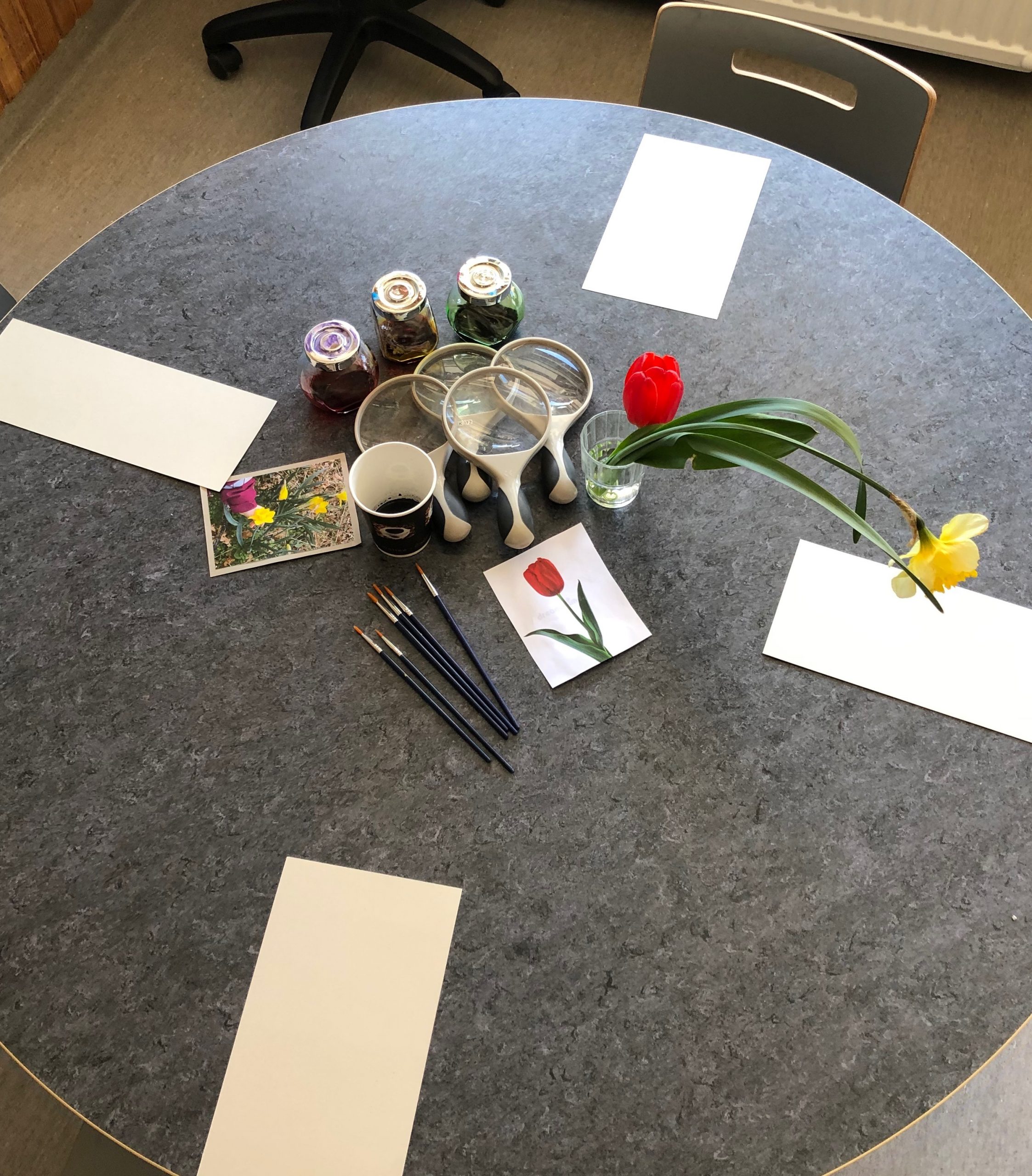 Ett bord med penslar och papper, vårtecken av blommor syns till.